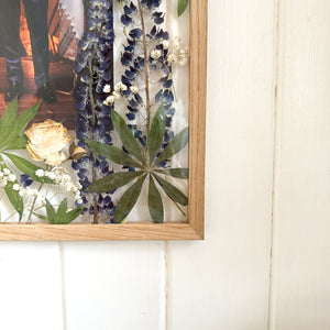 A close up of handcrafted pressed flower frame featuring a couple's wedding photo, vibrant lupins and pink roses, perfect for home decor.