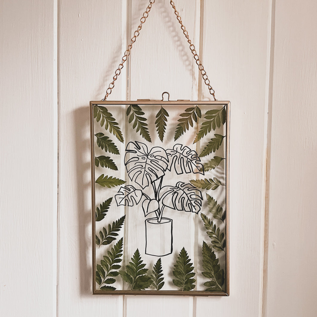 Handcrafted pressed flower frame featuring hand drawn Monstera Leaf. With fern leaves circling the drawing in a gold glass frame, perfect for home decor